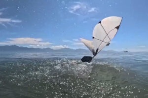 White flapping-wing aerial-aquatic robot emerges from the ocean, spraying water against a backdrop of mountains and blue sky.